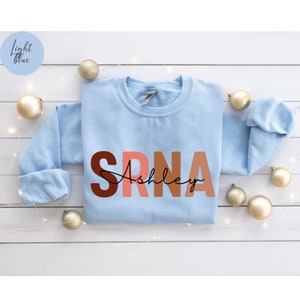 May include: Light blue sweatshirt with the letters "SRNA" in a gradient of brown, pink, and orange. The name "Ashley" is written in cursive below the letters.