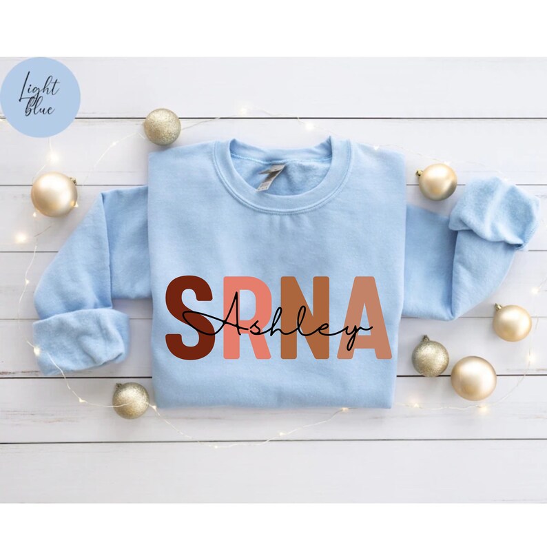 May include: Light blue sweatshirt with the letters "SRNA" in a gradient of brown, pink, and orange. The name "Ashley" is written in cursive below the letters.