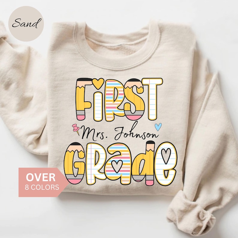 Puede incluir: Sudadera de color arena con las palabras "FIRST Mrs. Johnson GRADE" en un dise&ntilde;o colorido con tem&aacute;tica de l&aacute;pices. El texto est&aacute; en una fuente l&uacute;dica con corazones y detalles de l&aacute;pices. La sudadera tambi&eacute;n tiene el texto "OVER 8 COLORS".
