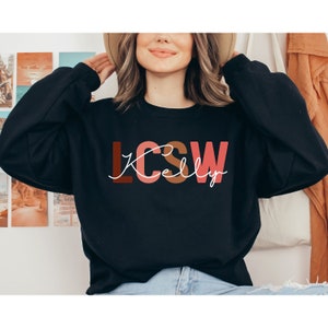 K&ouml;nnte beinhalten: Schwarzer Pullover mit dem Text "LCSW Kelly" in bunten Blockbuchstaben. Die Buchstaben sind in den Farben Rosa, Braun und Rot gehalten.