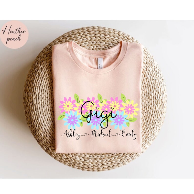 May include: A peach colored t-shirt with a floral design and the word "Gigi" in black script. Below the floral design, the names "Ashley", "Michael", and "Emily" are written in black script.