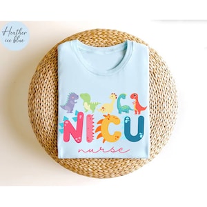 Pu&ograve; includere: T-shirt azzurra con scollo tondo, con un design di dinosauri colorati sopra la parola "NICU" in grandi lettere multicolore e la parola "nurse" in corsivo. La maglietta &egrave; piegata su una superficie circolare intrecciata.