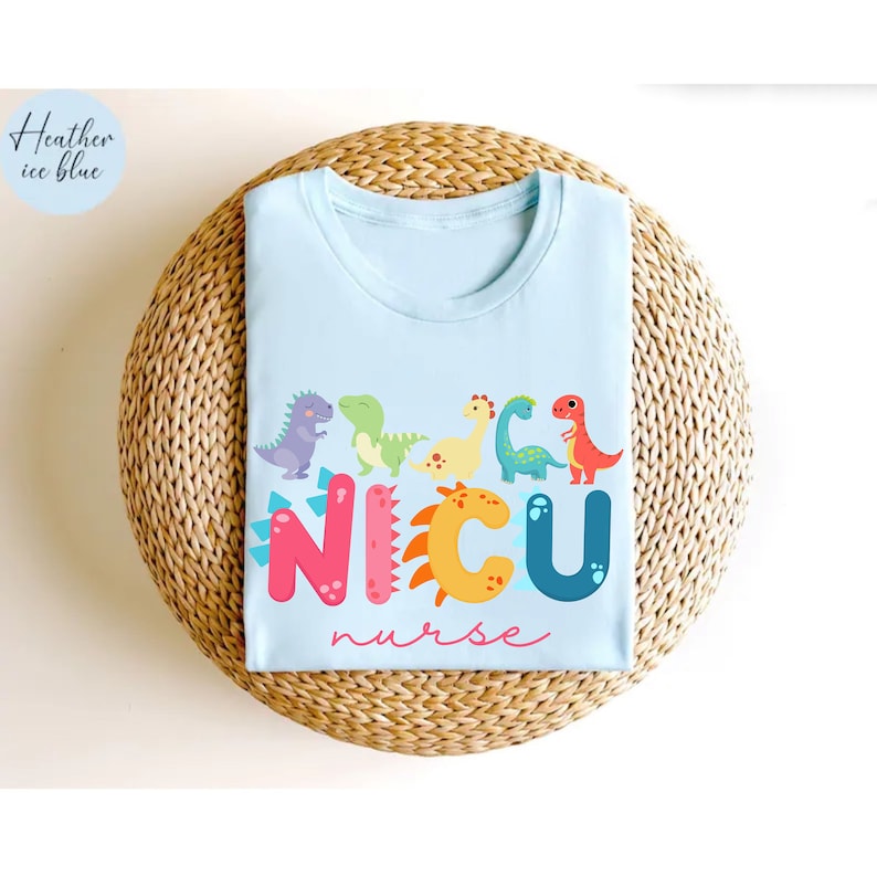Pu&ograve; includere: T-shirt azzurra con scollo tondo, con un design di dinosauri colorati sopra la parola "NICU" in grandi lettere multicolore e la parola "nurse" in corsivo. La maglietta &egrave; piegata su una superficie circolare intrecciata.