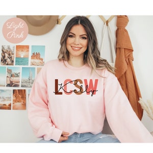 K&ouml;nnte beinhalten: Ein hellrosa Sweatshirt mit dem Text "LCSW Kelly" in braunen, rosa und roten Buchstaben.
