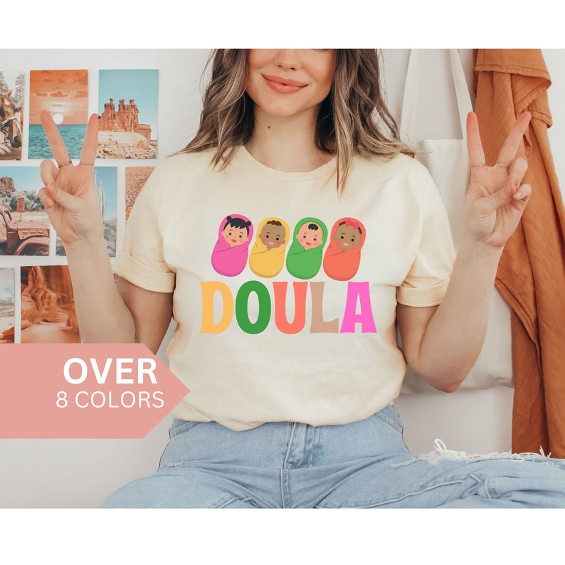 May include: A light yellow t-shirt with a graphic of four babies wrapped in blankets in different colors. The text "DOULA" is printed below the babies in rainbow colors.