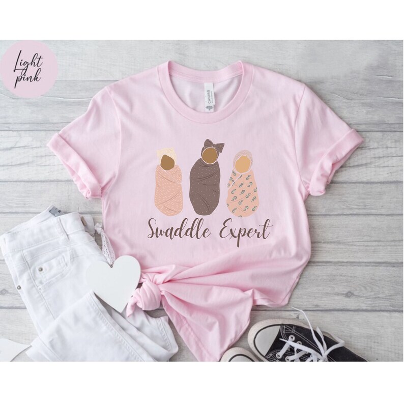 May include: A pink t-shirt with the text "Swaddle Expert" and a graphic of three babies swaddled in different patterns.