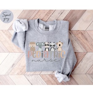 Puede incluir: Una sudadera gris con el texto "Pediatric Nurse" en una fuente colorida. El texto est&aacute; rodeado de cinco animales de dibujos animados: una oveja, un elefante, un mapache, un koala y un canguro.
