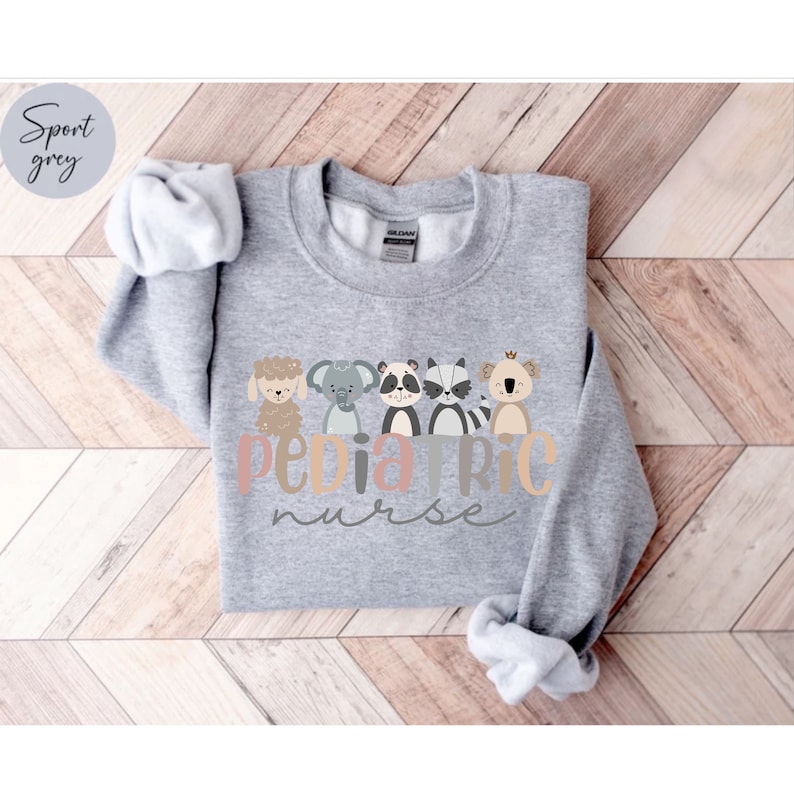 Puede incluir: Una sudadera gris con el texto "Pediatric Nurse" en una fuente colorida. El texto est&aacute; rodeado de cinco animales de dibujos animados: una oveja, un elefante, un mapache, un koala y un canguro.