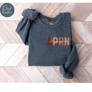 May include: A dark heather gray sweatshirt with the word 'APRN' printed in colorful letters. The letters are in shades of brown, orange, and pink. The word 'Jennifer' is written in cursive script underneath the letters.