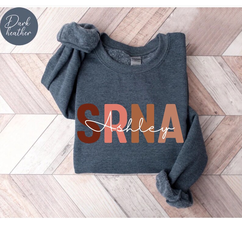 May include: Dark heather grey sweatshirt with the letters "SRNA" in brown, pink, and orange, with the name "Ashley" written in white cursive underneath.