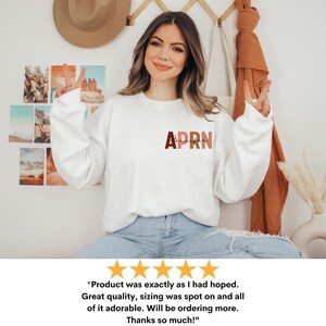 May include: A white sweatshirt with the text 'APRN' in brown, pink, and orange letters. The text is in a block font style. The sweatshirt is being worn by a person.