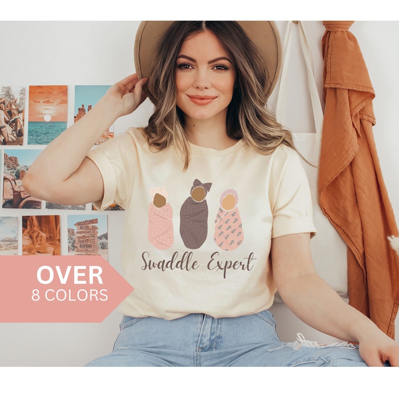 May include: A woman wearing a light beige t-shirt with a graphic of three swaddled babies. The text "Swaddle Expert" is printed below the graphic.