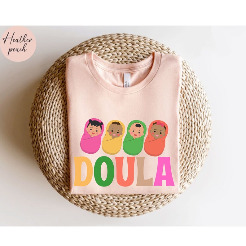 May include: A pink t-shirt with a graphic of four babies wrapped in blankets and the word "DOULA" in colorful letters.