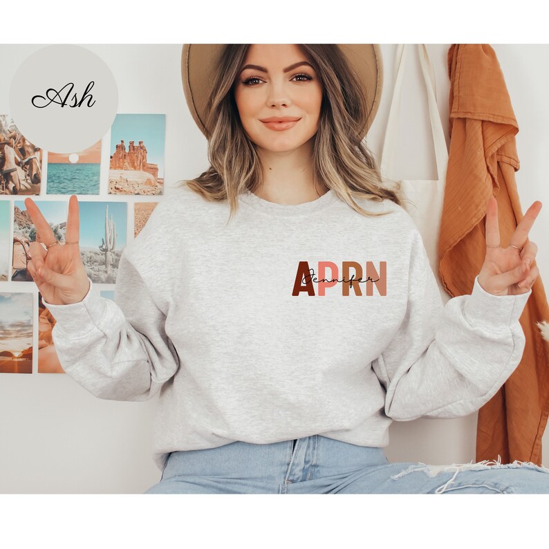 May include: A gray sweatshirt with the letters APRN in brown, pink, and orange colors. The letters are stacked on top of each other and the word 'Jennifer' is written in cursive underneath the letters.