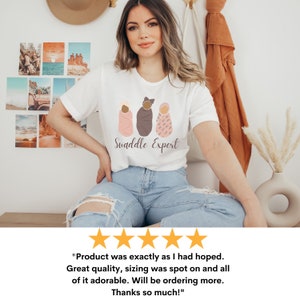 May include: A white t-shirt featuring a graphic of three swaddled babies in different skin tones. The text "Swaddle Expert" is printed below the graphic.