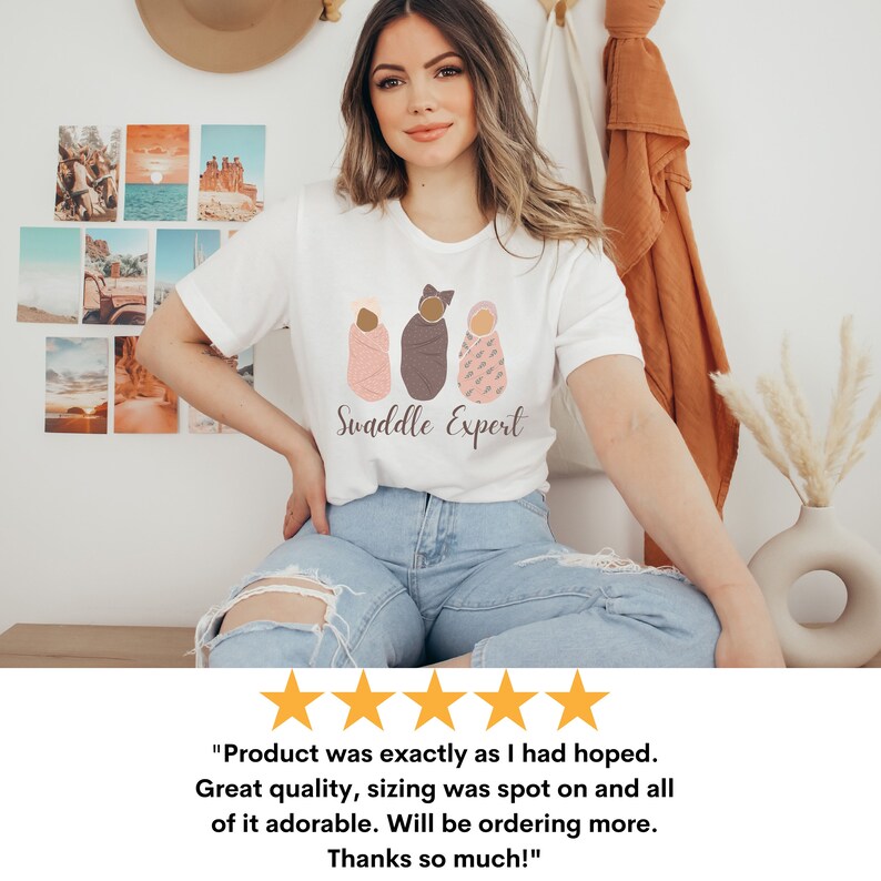 May include: A white t-shirt featuring a graphic of three swaddled babies in different skin tones. The text "Swaddle Expert" is printed below the graphic.
