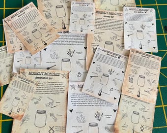 Op de afbeelding: Een verzameling verouderde papieren vellen met illustraties en tekst. De vellen bevatten tekeningen van potten, kaarsen en kruiden, met tekst als "MOONLIT MONTHLY" en titels als "money jar" en "love spell jar". De achtergrond is een groene snijmat.