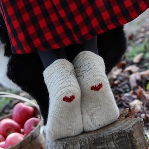 May include: Cream-colored knitted socks with red accents on the heels. A black and white cat is near a person wearing a red and black plaid skirt. A basket of red apples is in the foreground.