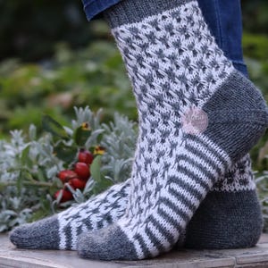 Könnte beinhalten: Ein Paar handgestrickte Wollsocken in Dunkelanthrazit und Weiß. Das Design zeigt ein komplexes Strukturmuster am Bein und Fuß, ergänzt durch einfarbige dunkelanthrazitfarbene Fersen, Zehenbereiche und Bündchen sowie horizontale Streifen.