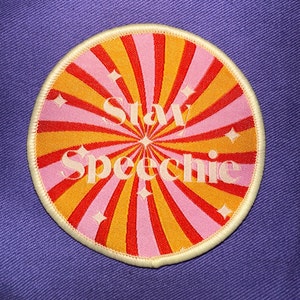 May include: A round embroidered patch with a retro design. The patch features a radiating pattern of pink, orange, and red stripes, with the words "Stay Speechic" in a light pink script. Small white star shapes are scattered throughout the design.