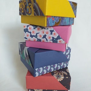Peut inclure: Une pile de quatre boîtes en origami avec du papier à motifs différents. La boîte du haut est jaune, la deuxième est rose avec des fleurs blanches, la troisième est bleue avec un motif blanc et or, et la boîte du bas est rouge avec un motif noir et or.