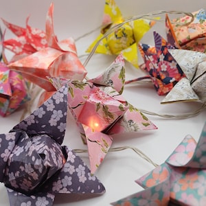 Peut inclure: Une guirlande lumineuse décorative avec des étoiles en origami. Les étoiles sont faites de papier à motifs dans des tons de rose, jaune et violet, avec des motifs floraux. Chaque étoile contient une petite lumière, reliée par un fil transparent.