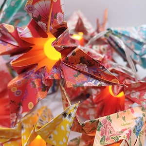 Pode incluir: Uma sequência de guindastes de papel origami com luzes internas. Os guindastes são feitos de papel estampado em vermelho, amarelo, laranja e azul-petróleo. Os padrões de papel incluem desenhos florais e de leques. As luzes são de cor amarelo quente.
