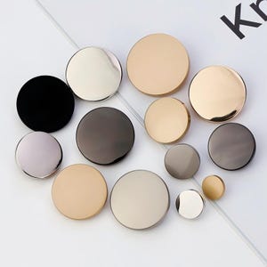 May include: Assortment of round metal buttons in various colors, including gold, silver, black, and gray. The buttons are arranged on a white surface.