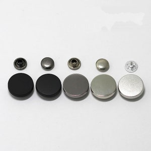 May include: Assortment of metal snap buttons in various finishes, including black, silver, and gold. The buttons are arranged in a row on a white surface.