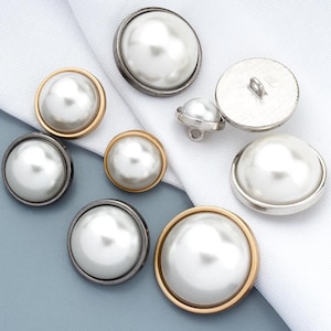 Op de afbeelding: Een verzameling ronde knopen met gladde witte kunstparelcentra in metalen randen. De knopen variëren in grootte en metaalafwerking, waaronder goud-, zilver- en donkere gunmetal-tinten. Elke knoop heeft een oogje aan de achterkant voor bevestiging.