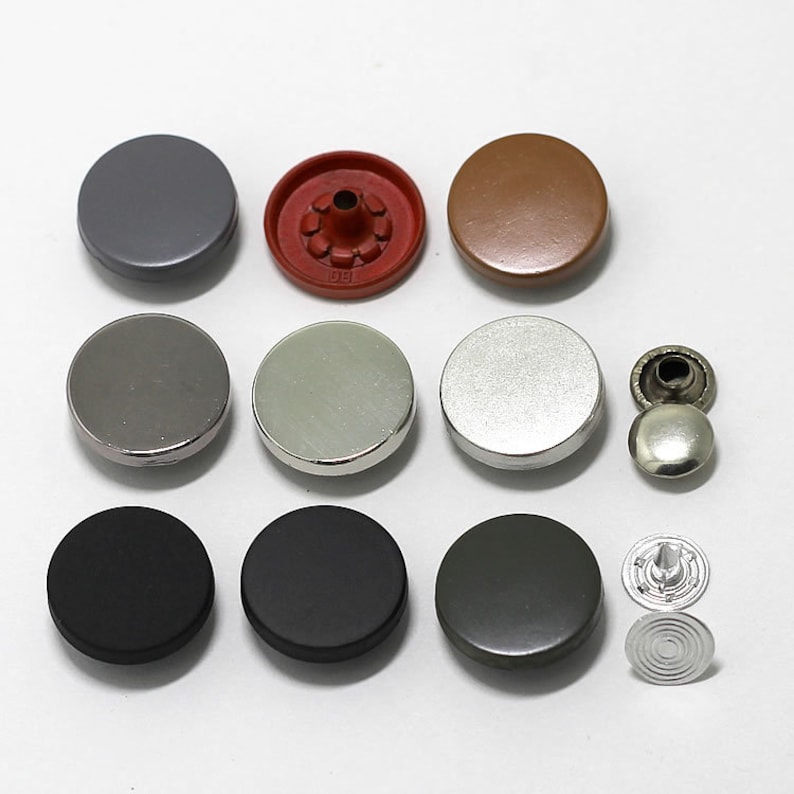 May include: Assortment of round metal buttons in various colors, including silver, black, gray, brown, and red. The buttons have a flat, smooth surface and a raised center.