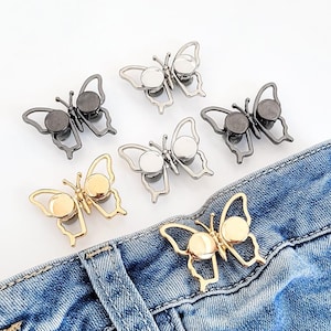 May include: A collection of six butterfly-shaped metal fasteners in silver, gold, and dark gunmetal tones. These decorative clips feature open-wing designs and are shown both loose and attached to the waistband of a pair of blue denim jeans.