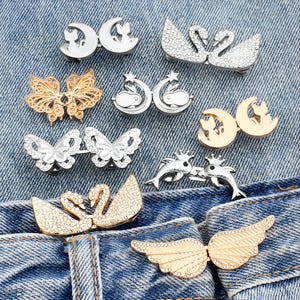 Op de afbeelding: Assortiment van zilveren en gouden metalen versieringen, waaronder vlinders, zwanen en vleugels. De versieringen zijn gerangschikt op een blauwe denim achtergrond.