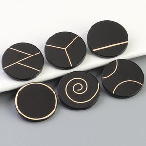 May include: Six black round buttons with gold geometric designs. The designs include a spiral, a circle with a line through it, a circle with two lines through it, a circle with three lines through it, a circle with a zig-zag line through it, and a circle with a straight line through it.