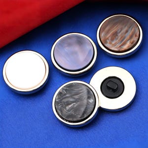 May include: A collection of five round buttons with metallic silver rims. The button faces vary in color and texture, including white, pearlescent purple, marbled brown, and black and white. One button has a black center.