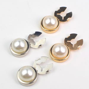 Pode incluir: Vários botões com efeito de pérola, com bordas de metal dourado e prateado e fechos correspondentes, espalhados sobre uma superfície branca. Os botões têm um design clássico e elegante, adequados para vários projetos de costura e artesanato.