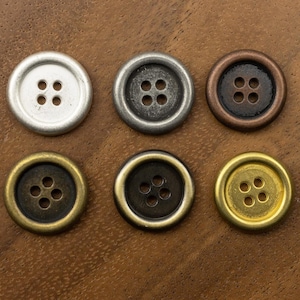 Op de afbeelding: Zes ronde knopen in verschillende metallic afwerkingen: zilver, tin, brons, messing en goud. Elke knoop heeft vier gaten om te naaien. De knopen zijn gerangschikt op een bruin houten oppervlak.