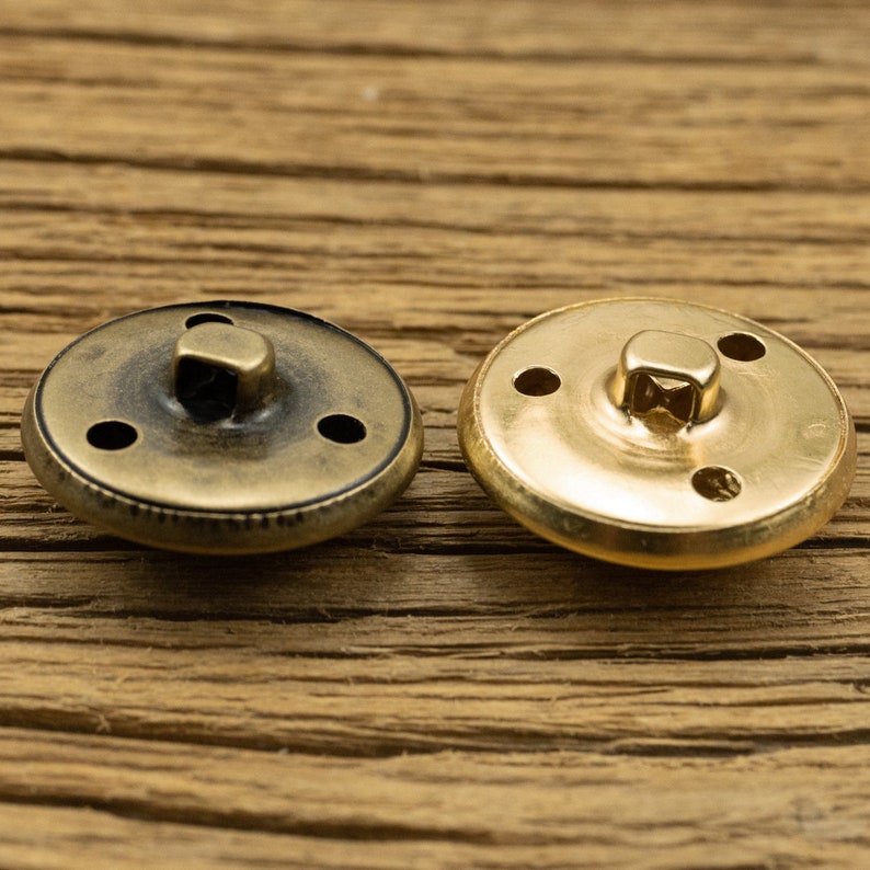 K&ouml;nnte beinhalten: Zwei Metallkn&ouml;pfe mit goldener Oberfl&auml;che. Ein Knopf hat eine dunklere, antike Oberfl&auml;che und der andere eine gl&auml;nzende, polierte Oberfl&auml;che. Beide Kn&ouml;pfe haben vier L&ouml;cher zum N&auml;hen.