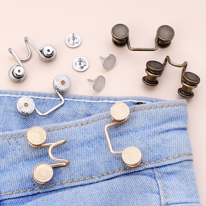 May include: A set of gold, silver, and bronze metal jean button extenders. The extenders are designed to add an extra inch to the waist of jeans. The extenders are shown on a pair of blue jeans.