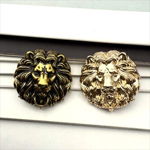 Puede incluir: Dos botones con cabeza de león, uno dorado y otro de bronce. Los botones son redondos y tienen un diseño detallado de una cabeza de león con melena.