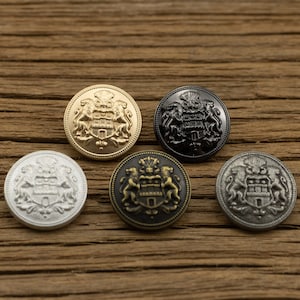 Puede incluir: Cinco botones decorativos con un diseño de escudo de armas. Los botones son de plata, oro, negro y bronce.