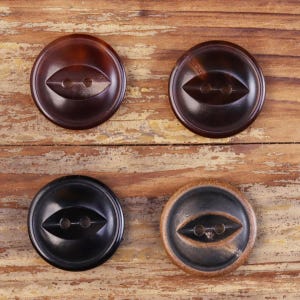 Puede incluir: Cuatro botones circulares dispuestos en formación cuadrada. Cada botón presenta una superficie lisa y pulida con una hendidura central en forma de ojo que contiene dos orificios para coser. Los colores incluyen marrón oscuro, negro y beige.