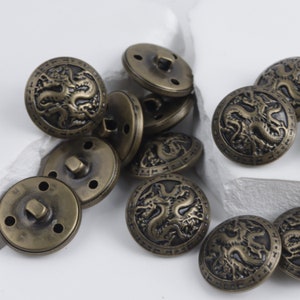 K&ouml;nnte beinhalten: Eine Gruppe antiker goldfarbener Kn&ouml;pfe mit einem Drachenmotiv. Die Kn&ouml;pfe haben ein erhabenes Design und sind aus Metall.