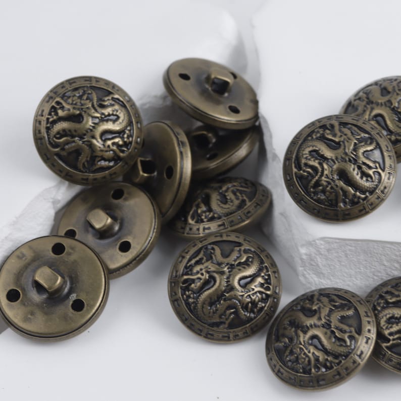 K&ouml;nnte beinhalten: Eine Gruppe antiker goldfarbener Kn&ouml;pfe mit einem Drachenmotiv. Die Kn&ouml;pfe haben ein erhabenes Design und sind aus Metall.