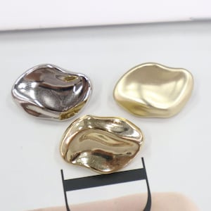 Könnte beinhalten: Drei unregelmäßig geformte Metallanhänger in Silber, Gold und Messing. Die Anhänger haben eine glatte, reflektierende Oberfläche und eine leicht konkave Form. Sie sind auf einer weißen Oberfläche angeordnet, was auf Schmuck- oder Bastelbedarf hindeutet.