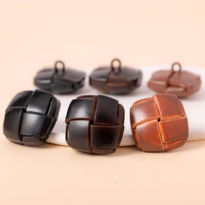 May include: Six leather buttons in various shades of brown and black. The buttons have a woven design and a loop on the back. The buttons are arranged on a light-colored surface.