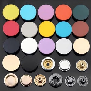 Puede incluir: Una colección de botones de presión metálicos circulares en varios colores sólidos, incluyendo amarillo, azul, lavanda, naranja, verde oscuro, rojo, blanco, negro y oro metálico. El set incluye tapas lisas y piezas de sujeción detalladas.