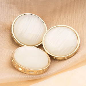 Może przedstawiać: Trzy białe okrągłe guziki ze złotą obwódką. Guziki mają perłowy połysk i subtelny wzór wirujący.