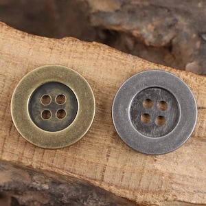 Könnte beinhalten: Zwei Metallknöpfe mit jeweils vier Löchern. Ein Knopf ist messingfarben, der andere silberfarben. Die Knöpfe sind rund mit einer leicht erhöhten Mitte. Die Knöpfe liegen auf einer Holzoberfläche.