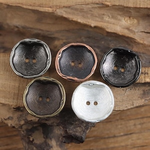 May include: Five round buttons with two center holes and a textured, organic surface pattern. The set includes one silver, one bronze, one copper-rimmed black, one dark gunmetal, and one black button, each featuring a slightly irregular, raised edge.